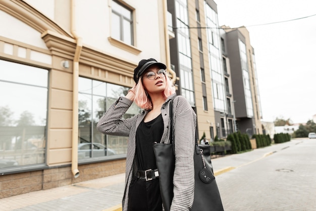 Urbana elegante bella giovane donna in occhiali vintage e un berretto in una camicia a quadri alla moda con una maglietta nera, jeans e una borsa alla moda passeggiate in città vicino all'edificio