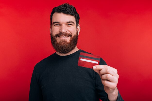 Usa la carta di credito, è più facile e intelligente Il giovane studente con la barba mostra una carta di credito, sorride sul muro rosso.