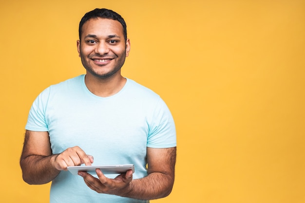 Usando la tavoletta. Felice vincitore! Giovane bell'uomo indiano afroamericano sorridente che tiene tablet e gioca o utilizza un'app di prenotazione isolata su sfondo giallo.