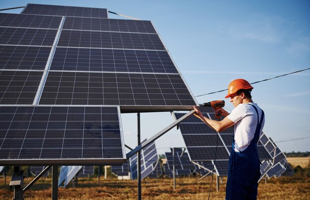 Usando un cacciavite a batteria. Lavoratore maschio in uniforme blu all'aperto con batterie solari al giorno pieno di sole.