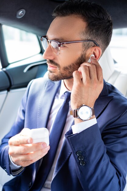 Uso degli auricolari. Bel giovane uomo d'affari barbuto che usa gli auricolari mentre è seduto in macchina