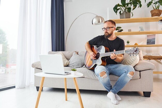 Uso del laptop L'uomo in abiti casual e con la chitarra acustica è al chiuso