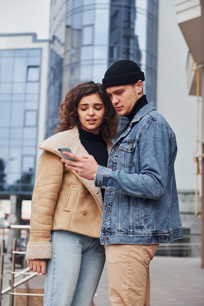 Uso del telefono Una coppia allegra in abiti caldi e casual fa una passeggiata all'aperto nella città vicino all'edificio degli affari