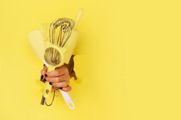 Utensili della cucina della tenuta della mano della donna su fondo giallo.