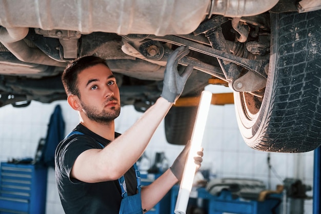 Utilizza apparecchiature di illuminazione L'uomo in uniforme da lavoro ripara l'automobile bianca all'interno Concezione del servizio automobilistico