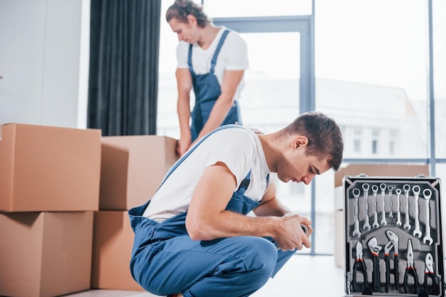 Utilizza gli strumenti della custodia Due giovani traslocatori in uniforme blu che lavorano all'interno della stanza