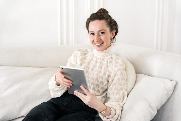 Utilizza un tablet appoggiato su un divano lavoro quotidiano in ufficio una giovane donna freelance sorridente camicia bianca