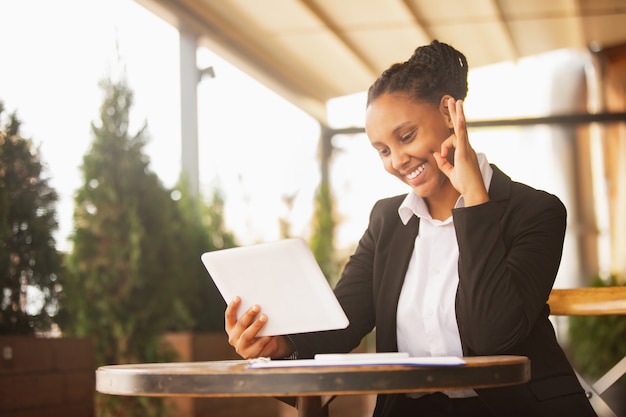 Utilizzando tablet, videochiamata. Donna d'affari afro-americana in abbigliamento da ufficio sorridente, sembra sicura di sé felice, impegnata. Concetto di finanza, affari, uguaglianza e diritti umani. Bella giovane modella, di successo.