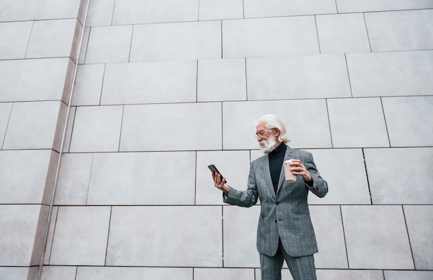 Utilizzo dello smartphone L'uomo d'affari senior in abiti formali con capelli grigi e barba è all'aperto