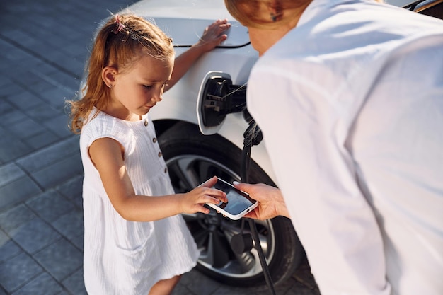 Utilizzo dello smartphone La giovane donna con la sua piccola figlia è con la loro auto elettrica all'aperto