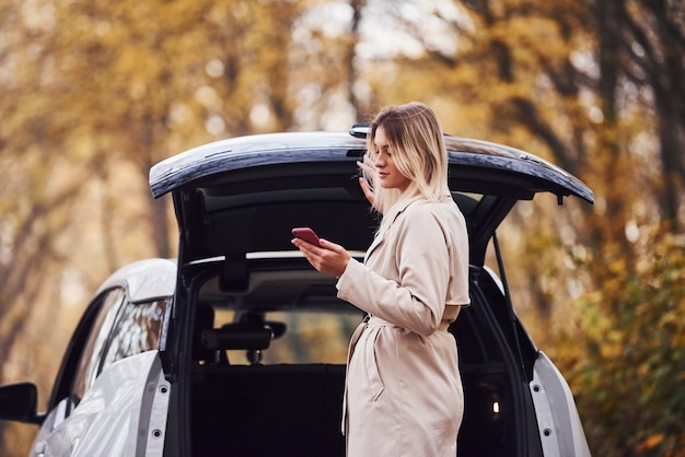 Utilizzo dello smartphone. La ragazza ha un viaggio autunnale in auto. Automobile nuova di zecca moderna nella foresta.