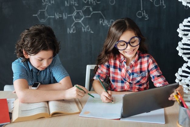 Utilizzo di gadget moderni per l'istruzione. Piccoli alunni felici positivi seduti a scuola e godersi la lezione di lingue straniere mentre prendono appunti e usano il dizionario