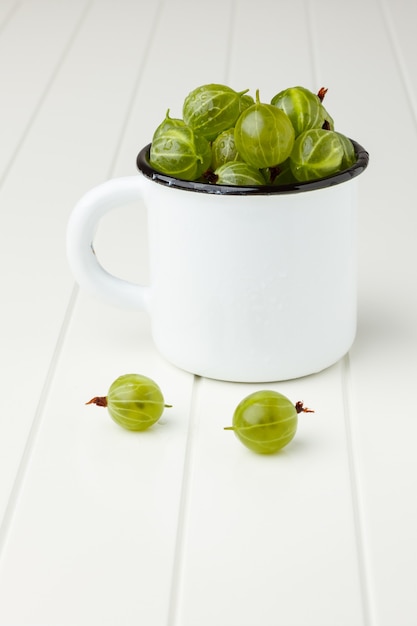 Uva spina verdi fresche in una tazza dello smalto sulla tavola bianca