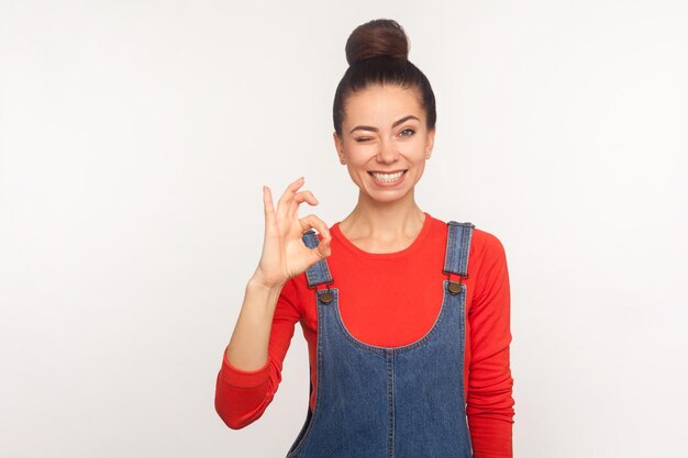 Va bene d'accordo con te Ritratto di ragazza carina ed elegante allegra con crocchia per capelli in tuta di jeans che mostra segno giusto della mano e ammiccante idea di approvazione indoor studio girato isolato su sfondo bianco