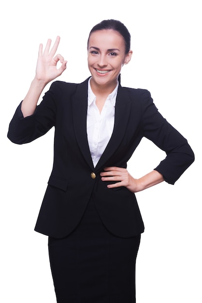 Va tutto bene! Felice giovane donna in abiti da cerimonia gesticolando segno OK e sorridendo in piedi isolato su bianco