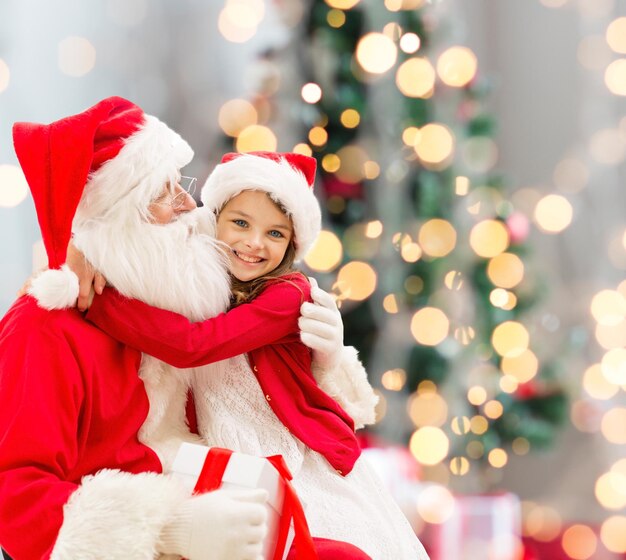 vacanze, celebrazione, infanzia e concetto di persone - bambina sorridente che abbraccia con babbo natale su sfondo di luci dell'albero di natale
