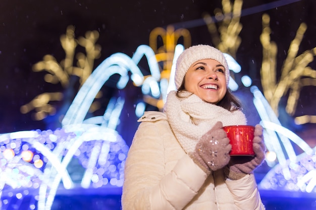 Vacanze di Natale. Bella donna sorridente in abiti caldi con una tazza di caffè caldo all'aperto in inverno. Notte della città.
