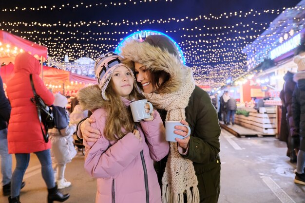 Vacanze di Natale e Capodanno, mamma felice e figlia bambino che camminano insieme bevendo una tazza di tè caldo al mercatino di Natale