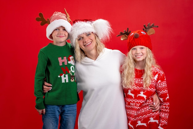 Vacanze di Natale Ritratto di una famiglia felice di tre persone In cappelli di Babbo Natale, madre e bambini in piedi e sorridente su sfondo rosso per studio