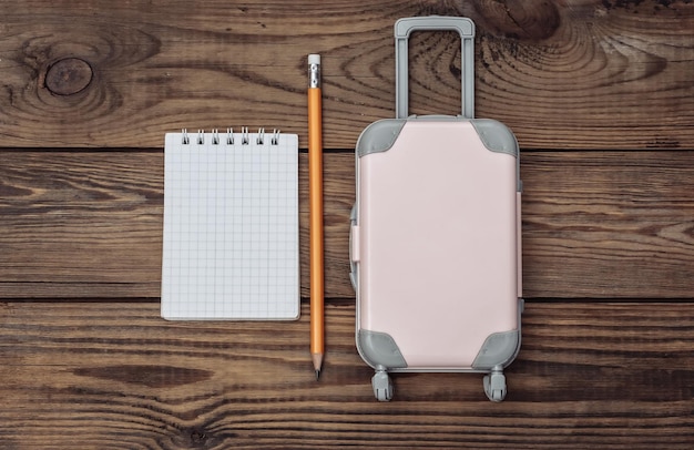 Vacanze di vacanza piatta e concetto di planata di viaggio. Mini valigia da viaggio in plastica e taccuino su fondo di legno. Vista dall'alto