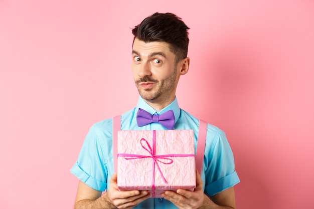 Vacanze e concetto di celebrazione. Ragazzo divertente con i baffi che mostra la confezione regalo carina avvolta in carta festosa, che guarda l'obbiettivo, che ti presenta, in piedi su sfondo rosa.