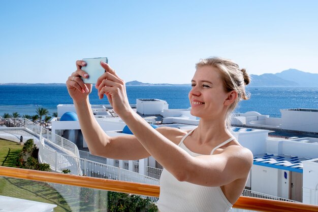 Vacanze e viaggi. Donna felice in costume da bagno che si fa selfie sul cellulare in piedi sul balcone della camera d'albergo, vista mare