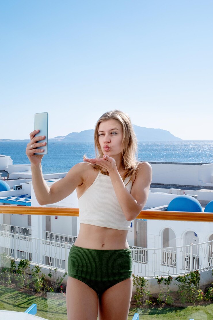 Vacanze e viaggi. Donna felice in costume da bagno che si fa selfie sul cellulare in piedi sul balcone della camera d'albergo, vista mare