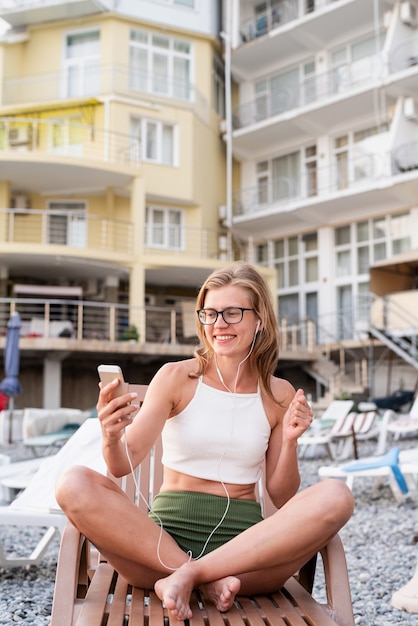 Vacanze estive sulla spiaggia. La bella giovane donna in costume da bagno seduta sul lettino che ascolta la musica utilizzando il dispositivo mobile e balla, ora blu