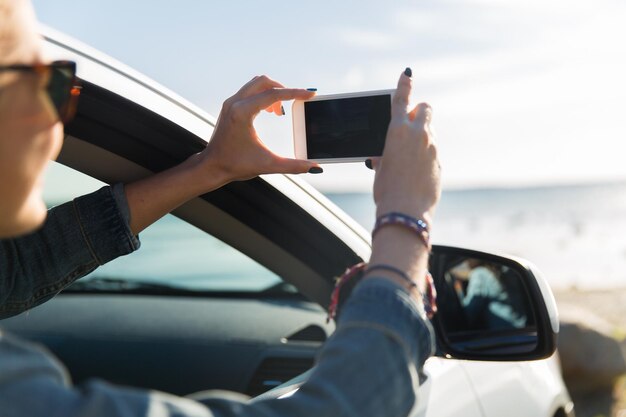 vacanze estive, vacanze, viaggi, viaggio su strada e concetto di persone - adolescente felice o giovane donna in auto che scatta foto del mare con lo smartphone