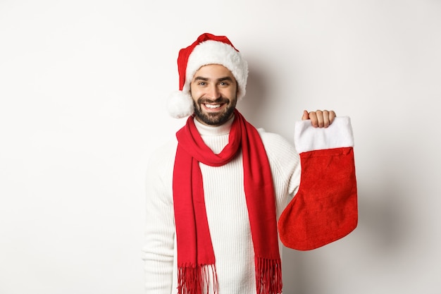 Vacanze invernali e concetto di celebrazione. Felice uomo barbuto con cappello da Babbo Natale che tiene in mano un calzino natalizio, portando regali, in piedi su sfondo bianco