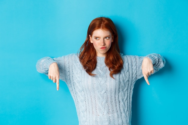 Vacanze invernali e concetto di persone. Ragazza rossa indecisa in maglione che punta le dita verso il basso e pensa, avendo dubbi, in piedi su sfondo blu.