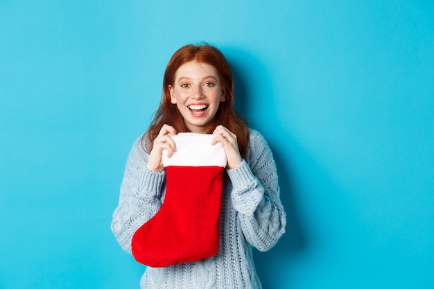 Vacanze invernali e concetto di regali. Felice ragazza dai capelli rossi che riceve un regalo di Natale, apre la calza di Natale e sorride stupita, in piedi su sfondo blu