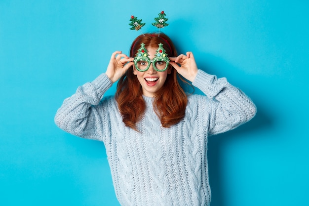 Vacanze invernali e concetto di vendite di Natale. Bellissima modella rossa che celebra il nuovo anno, indossa una divertente fascia per capelli e occhiali, sorridente sciocco, sfondo blu