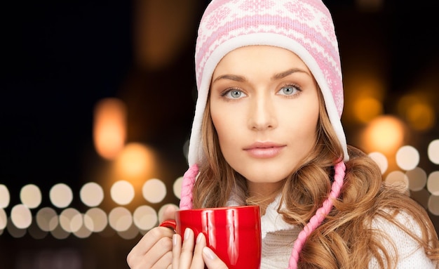 vacanze, Natale e concetto di persone - primo piano di una giovane donna con cappello d'inverno con una tazza rossa sullo sfondo delle luci