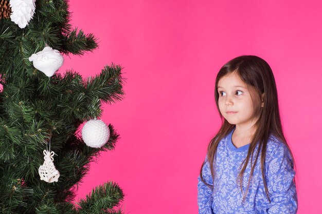 Vacanze per bambini e concetto di natale bambina che decora l'albero di natale su sfondo rosa