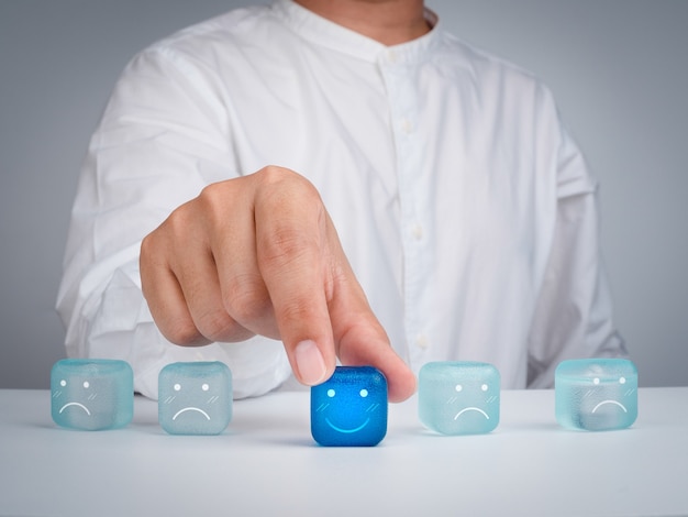 Valutazione del servizio clienti, feedback, valutazione, esperienza del cliente e concetti di indagine sulla soddisfazione. La mano dell'uomo d'affari sceglie l'icona del sorriso felice sul cubo blu sul tavolo e sullo sfondo bianco.