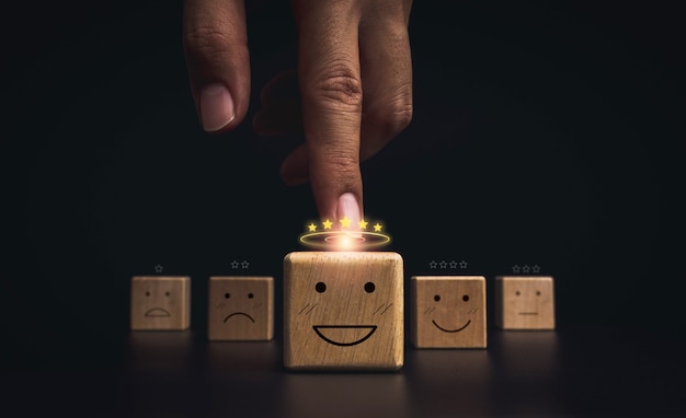 Valutazione del servizio clienti, valutazione, feedback e concetto di indagine sulla soddisfazione. Mano che indica sul viso di emoticon sorriso felice con cinque stelle dorate sul blocco di legno su sfondo scuro.