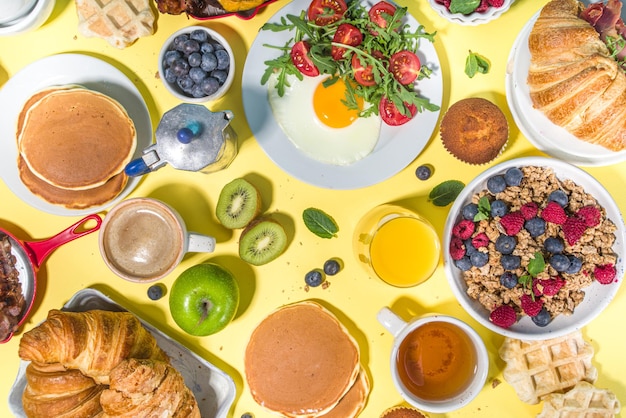 Vari cibi tradizionali per la colazione uova fritte con pancetta muesli avena cialde frittelle hamburger croissant frutta bacche caffè tè e succo d'arancia giallo tavolo sfondo spazio copia vista dall'alto