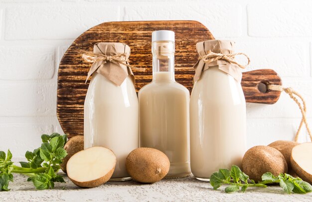 Vari tipi di bottiglie di latte di patate dal muro di mattoni bianchi sullo sfondo di una tavola da cucina in legno tuberi e foglie di patate nelle vicinanze