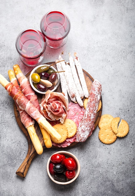 Varietà di affettati e antipasti, vino rosso, prosciutto, prosciutto, affettati, salsiccia, grissini, olive. Misto assortito di carne su tavola di legno rustica, sfondo di pietra grigia, vista dall'alto, primo piano