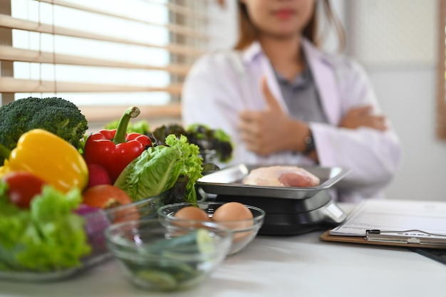 Varietà di verdure fresche in primo piano, carne e uova su tavolo bianco con un nutrizionista seduto sullo sfondo