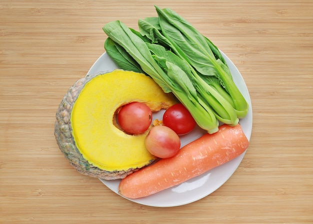 Varietà di verdure fresche sul piatto bianco contro il fondo del bordo di legno.