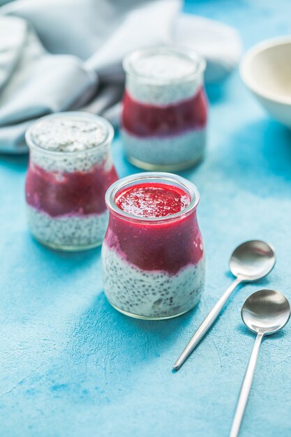 Vasetti di vetro con budino di chia con lampone e marmellata. Ciotola di bacche di lampone su sfondo blu.