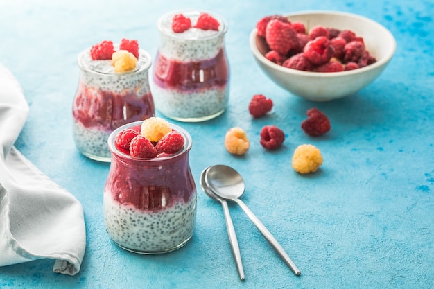 Vasetti di vetro con budino di chia con lampone e marmellata. Ciotola di bacche di lampone su sfondo blu.