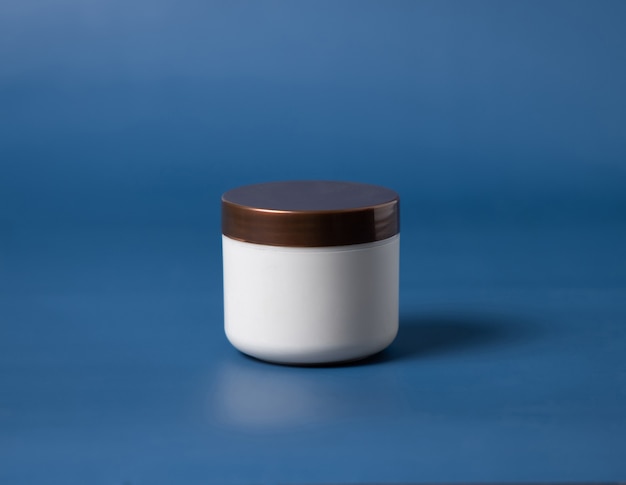 Vaso bianco crema con coperchio marrone su sfondo blu vicino. La crema cosmetica può mockup