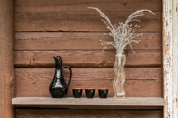 Vaso con fiori secchi e una brocca con bicchierini contro una parete di legno marrone