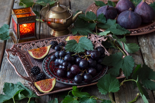 vaso di bronzo con uva nera, fichi, more, dietro un vecchio teiera e candelabro con candele