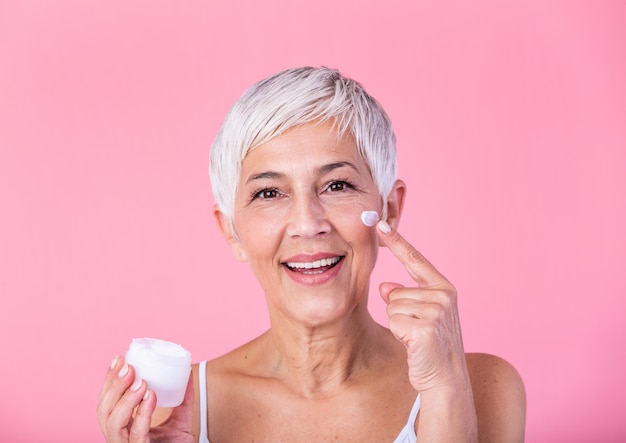 Vaso di detenzione bella donna matura di crema idratante e guardando la fotocamera. Donna senior felice che tiene una bottiglia di lozione anti-invecchiamento isolata sopra fondo rosa. Trattamento anti invecchiamento e bellezza.