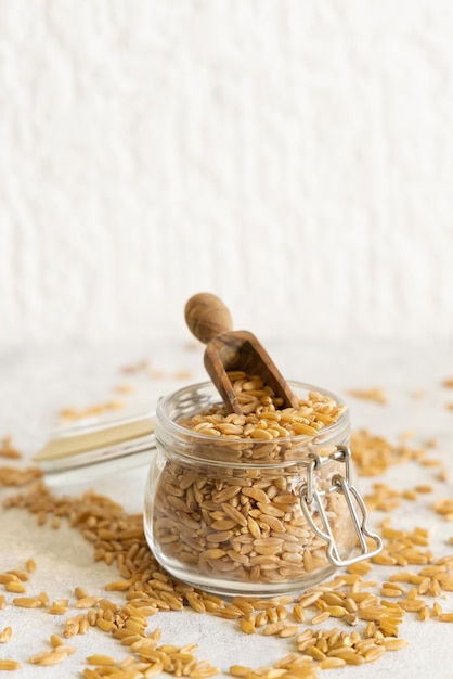 Vaso di vetro di grano di segale secco crudo con un cucchiaio di legno sulla tavola bianca da vicino