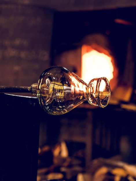 Vaso in vetro trasparente con tubo metallico per la soffiatura del vetro posizionato vicino al forno ardente e alla legna da ardere in un'officina professionale